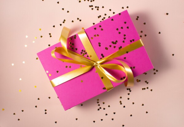 A bright pink gift box wrapped in gold ribbon is covered in shiny gold confetti.