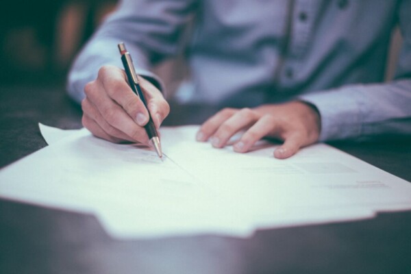 A man wearing a grey dress shirt holds a pen and signs documents.