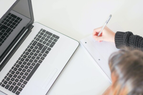 A woman holds a pen and write in a notebook with an open laptop in front of her